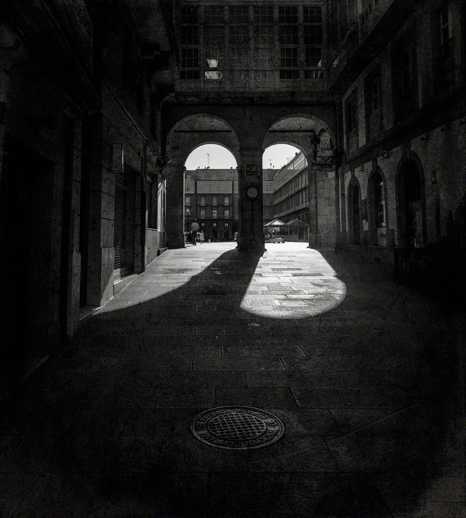 A narrow, dimly lit alleyway with arches, casting long shadows on the stone pavement, leading to a brighter square in the distance.