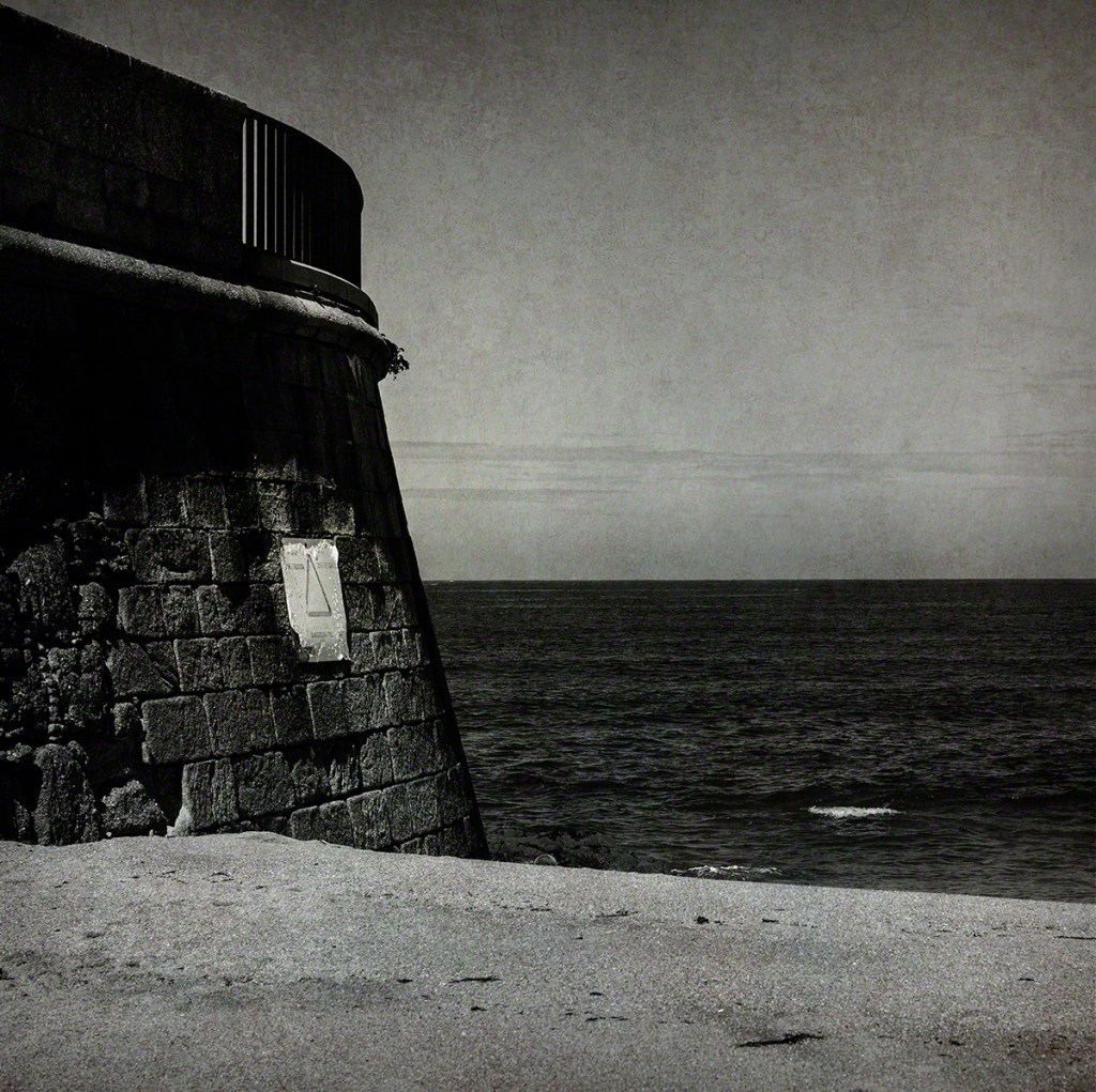 A black and white photograph of a stone pier extending into the ocean, with a partly cloudy sky above and rippling water visible in the background.