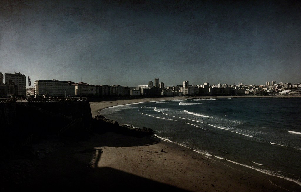 View of a beach and city skyline under a clear blue sky, with gentle waves lapping at the shore.