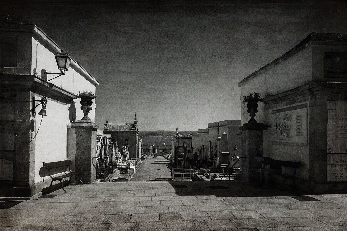 A desolate cemetery path lined with elaborate tombs and monuments, featuring a bench on the left and a solitary figure sitting nearby, under a clear sky.