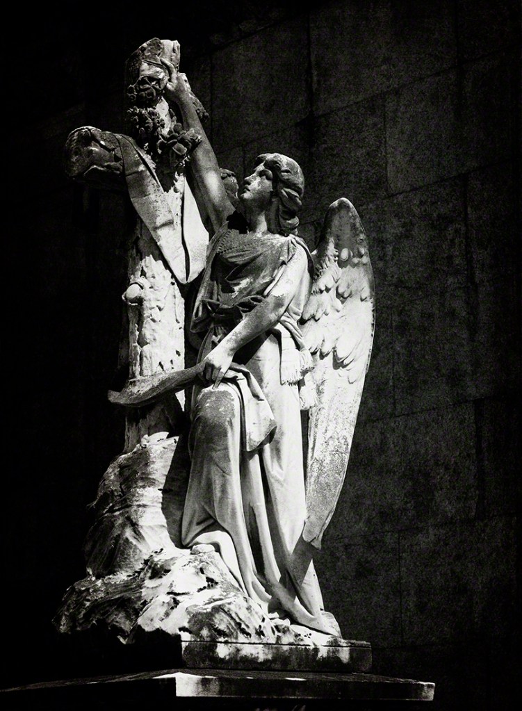Statue of an angel holding a sword, portrayed in a dramatic black and white setting beside a stone cross.