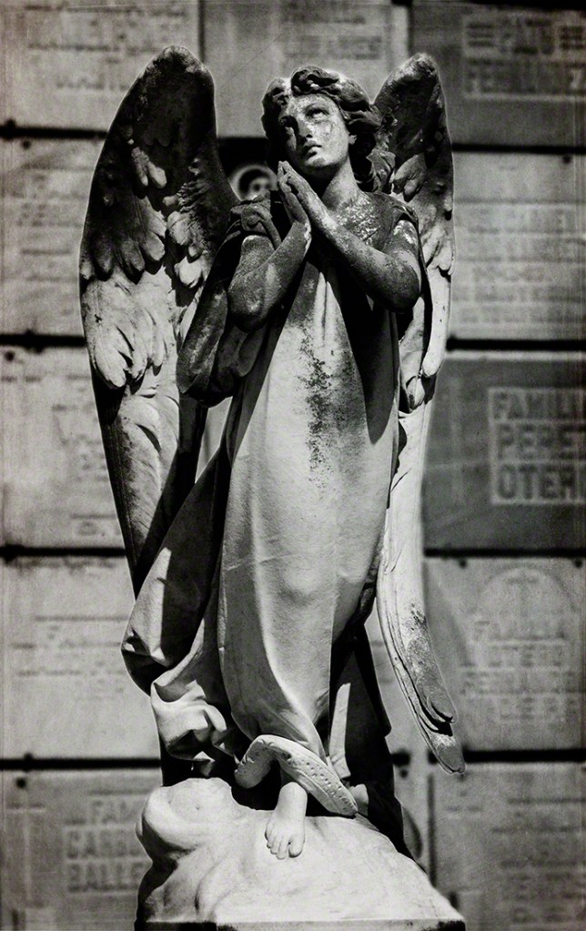 A black and white photograph of a weathered angel statue with wings, standing with hands clasped in prayer.
