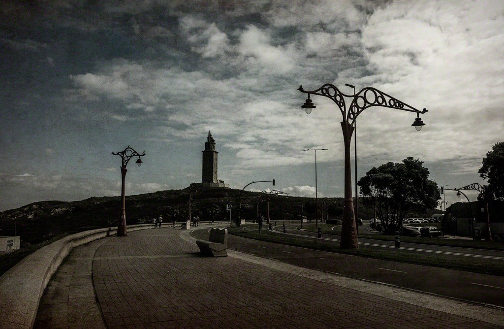 A scenic view of a coastal promenade with artistic street lamps, leading towards a lighthouse situated on a hill under a cloudy sky.