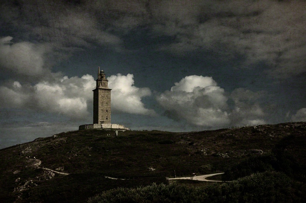 A historic lighthouse situated atop a grassy hill under a dramatic sky with clouds.