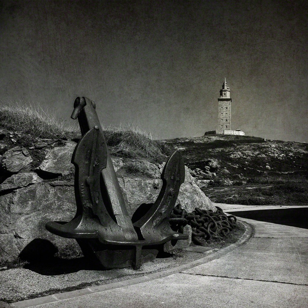 A black and white image featuring two large anchors in the foreground, positioned on a rocky surface with grass. In the background, a tall lighthouse stands on a hill against a cloudy sky.