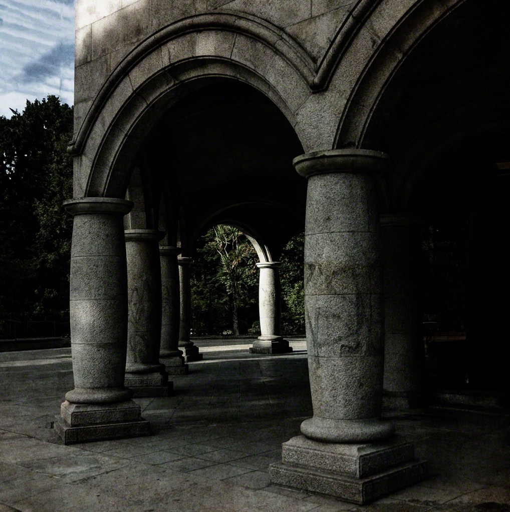 Stone pillars supporting arched structures in a shaded outdoor area, with greenery in the background.