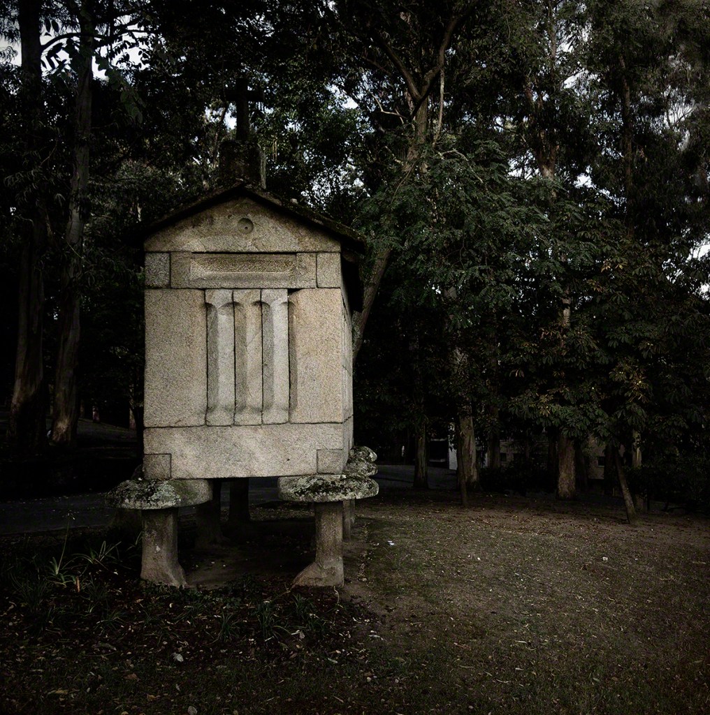 A stone structure resembling a small shrine or monument, with four vertical columns and a sloped roof, surrounded by trees in a park setting.